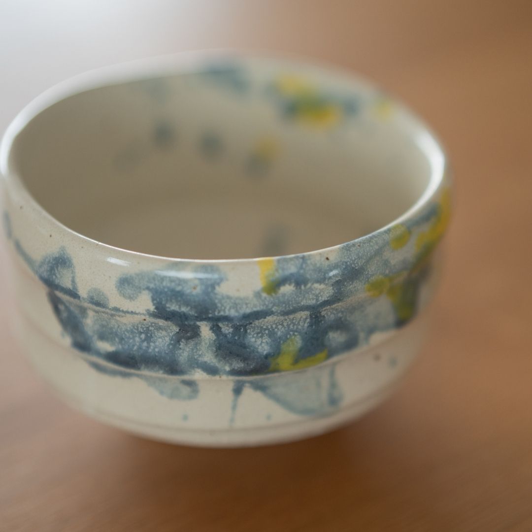 Draufsicht der Matcha-Schale "Splash" mit lebendigen blauen und gelben Glasurflecken auf weißem Shino-Ton, inspiriert von der kühlenden Tradition des Uchimizu im japanischen Sommer.
