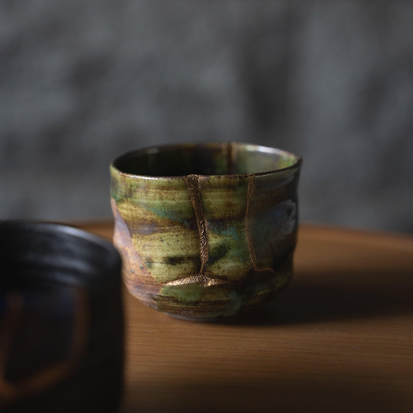 Dyb grøn te-skål repareret med Kintsugi, en traditionel japansk teknik, der viser skønheden i uperfekthed, placeret på et træbord i japansk stil.