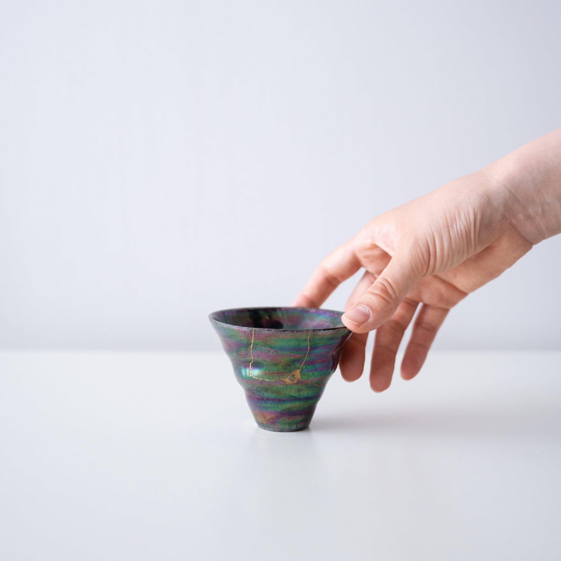 Hånd, der holder en iriserende koralformet kop repareret med Kintsugi, en traditionel japansk teknik.