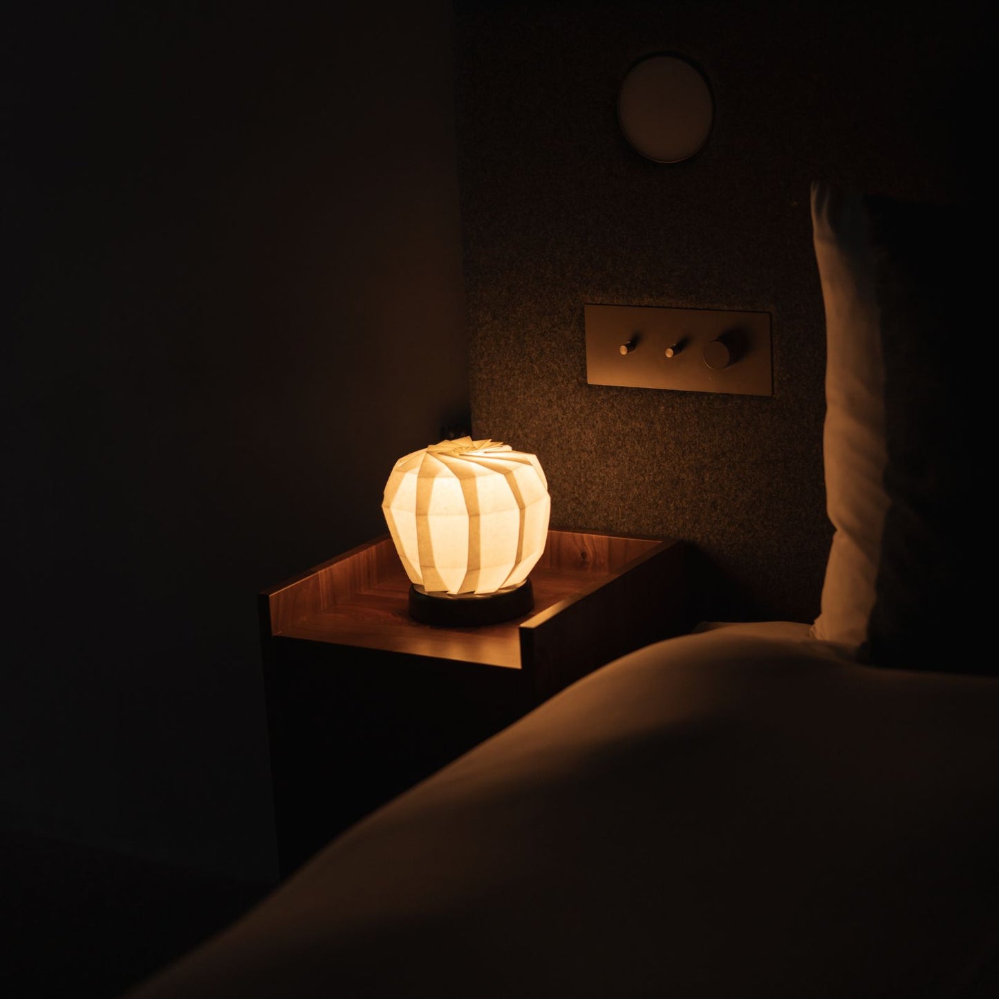 Japanse Origami Papieren Tafellamp "Bloem van Licht" - maru M die warm licht uitstraalt op een nachtkastje in een Japandi slaapkamer.