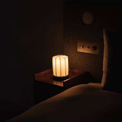 Japanse Origami Papieren Tafellamp "Bloem van Licht" - tutu M die warm licht uitstraalt op een nachtkastje in een Japandi slaapkamer.
