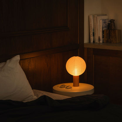 "Een schemerige slaapkamer met links een bed en rechts een boekenplank met boeken. Op het nachtkastje rechts van het bed staat een nachtlampje \"TORCHIN\" gemaakt van Japanse Yame-chochin lantaarns, dat een warme gloed uitstraalt vanuit de sferische kap."