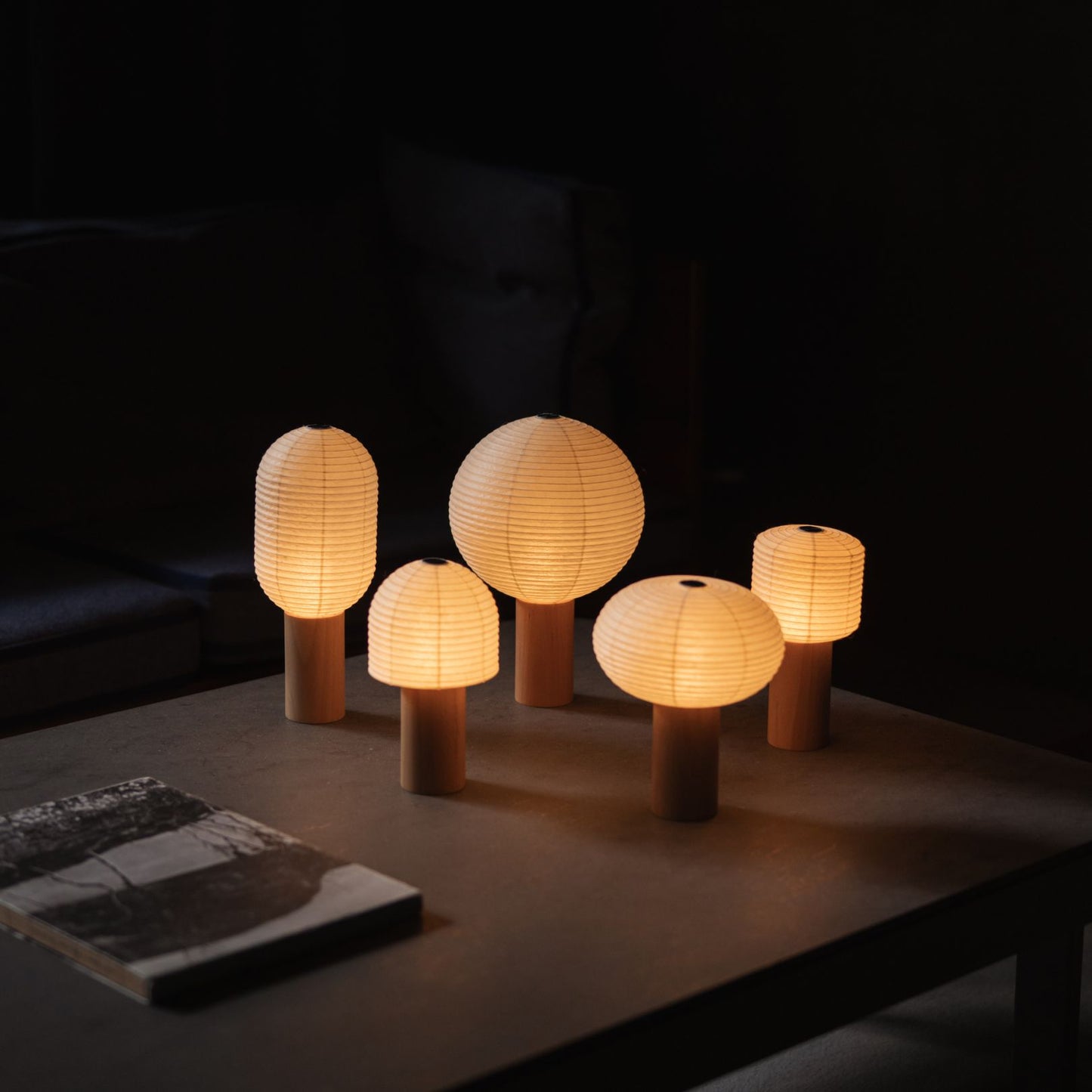 Fünf verschiedene japanische Papier-Tischlampen „TORCHIN“, die warmes Licht auf einen grauen Tisch in einem schwach beleuchteten Raum werfen.
