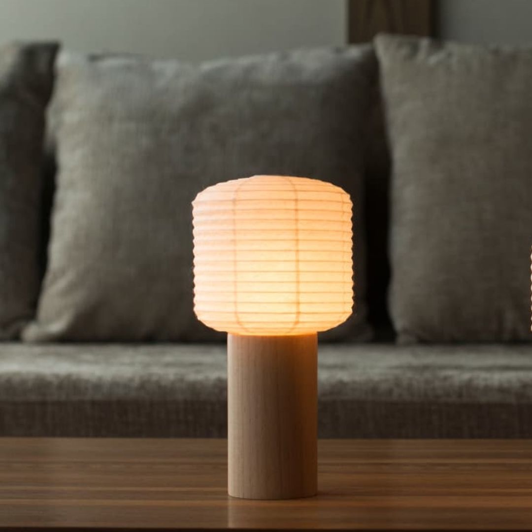 Seitenansichtsfoto einer tragbaren japanischen Papierlampe „Torchin“, die auf einem Holztisch steht. Im Hintergrund ist ein graues Sofa zu sehen. Der Lampenschirm der Torchin besteht aus einer japanischen Laterne mit einem darunter befestigten zylindrischen Holzgriff.