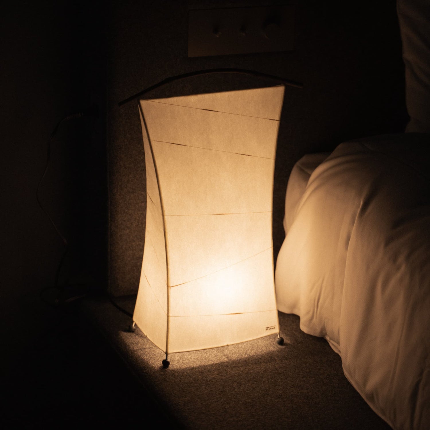 "Une lampe japonaise en papier diffuse une lumière chaude à côté d'un lit dans une chambre faiblement éclairée"