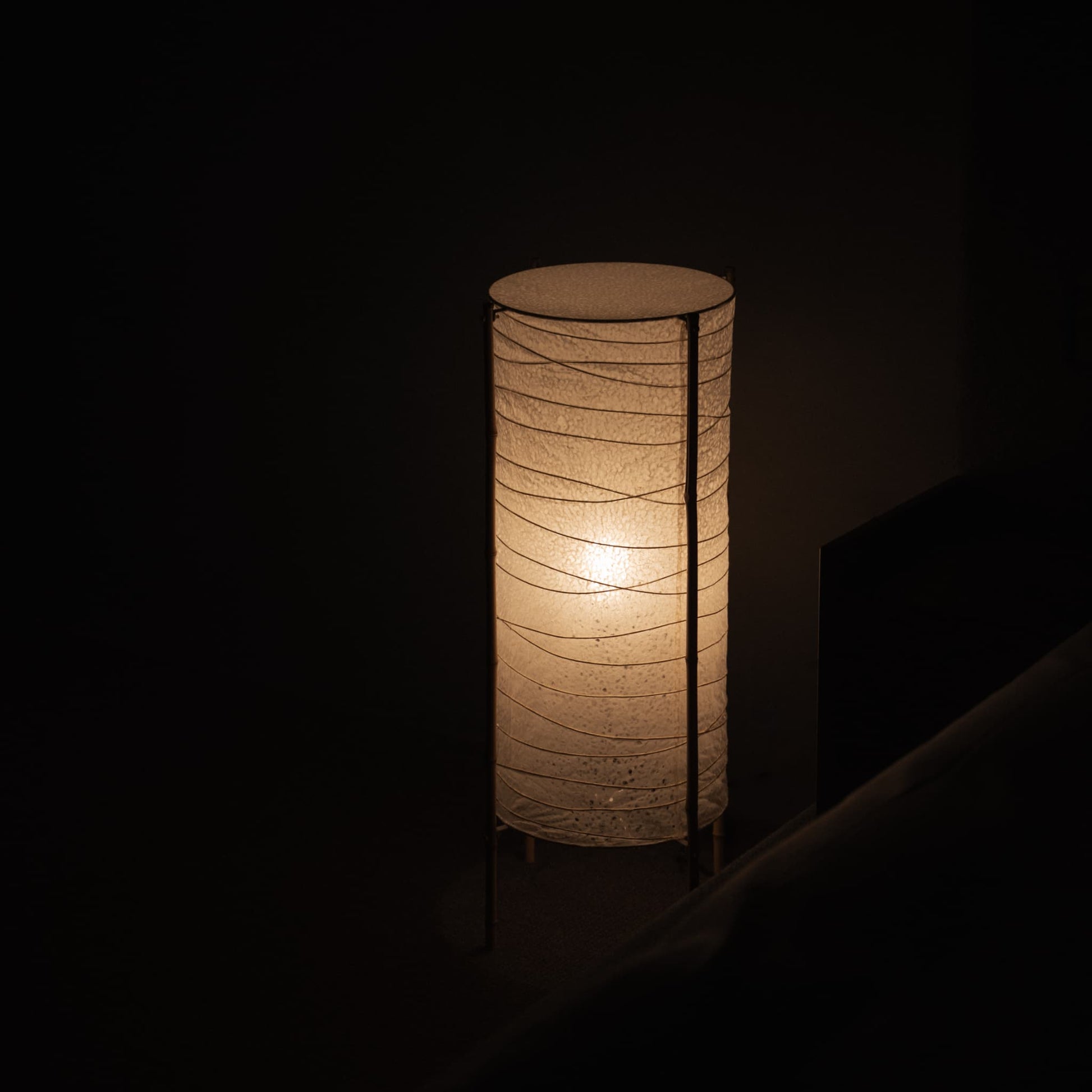 Salle faiblement éclairée avec un lampadaire japonais cylindrique en papier washi et un cadre en bambou, projetant une lueur chaleureuse dans une chambre japonaise