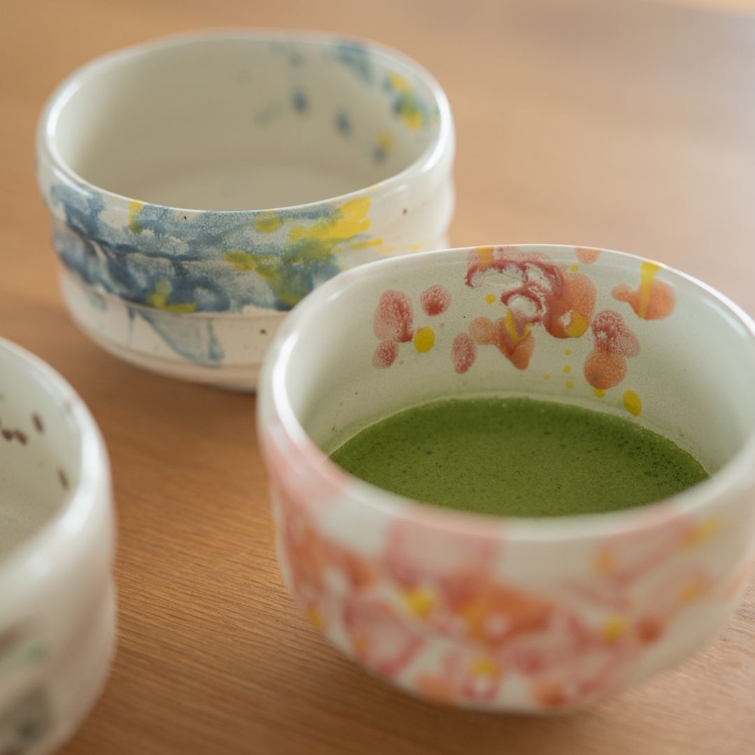 Vinklet billede af Shino-stellet med matcha-skåle på et træbord, med "Flower"-skålen med tilberedt grøn matcha-te og de to andre designs.