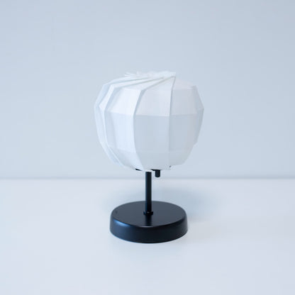 "Stijlvolle origami tafellamp met een zwarte standaard en een sferische witte lampenkap, vervaardigd uit ingewikkeld gevouwen washipapier."