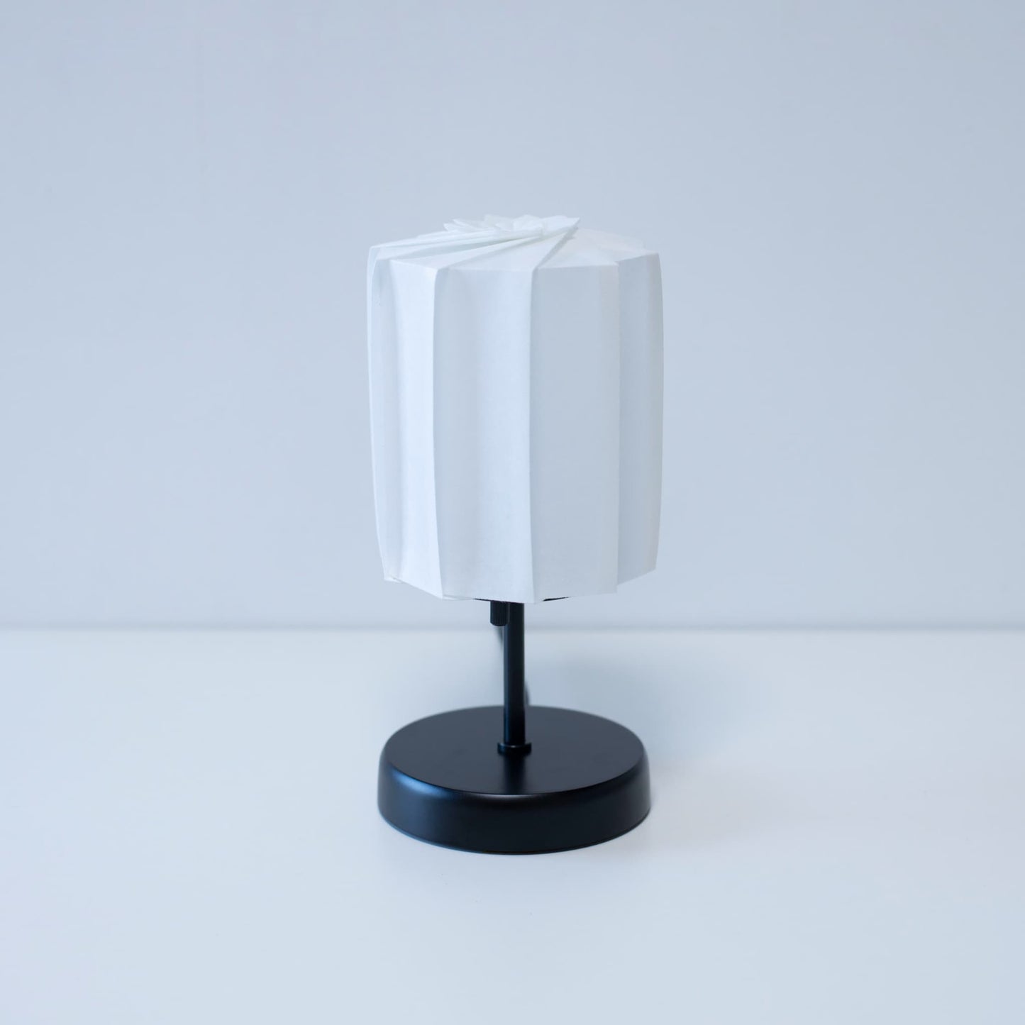 "Origami tafellamp \"tutu\" met een zwarte standaard en een witte cilindrische lampenkap van geplooid washipapier, wat een elegante toets geeft aan elke kamer."