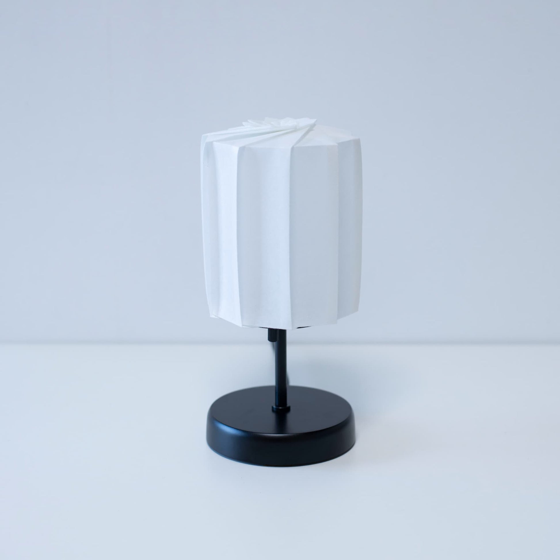 "Origami tafellamp \"tutu\" met een zwarte standaard en een witte cilindrische lampenkap van geplooid washipapier, wat een elegante toets geeft aan elke kamer."