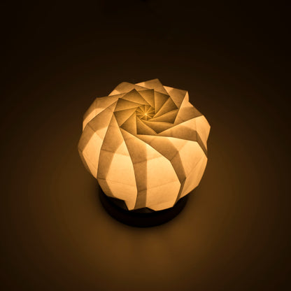 Origami papieren tafellamp met een zwarte voet en een bolvormige witte lampenkap van ingewikkeld gevouwen washipapier, die zacht en warm licht uitstraalt