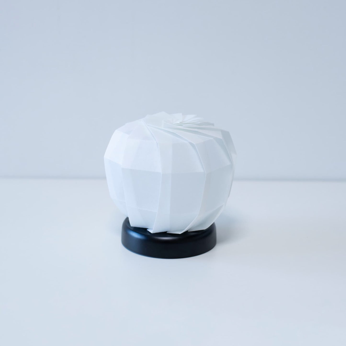 "Moderne Origami tafellamp \"maru M\" met een zwarte voet en een witte, getextureerde lampenkap, die zorgt voor een warme en uitnodigende sfeer in elke kamer."