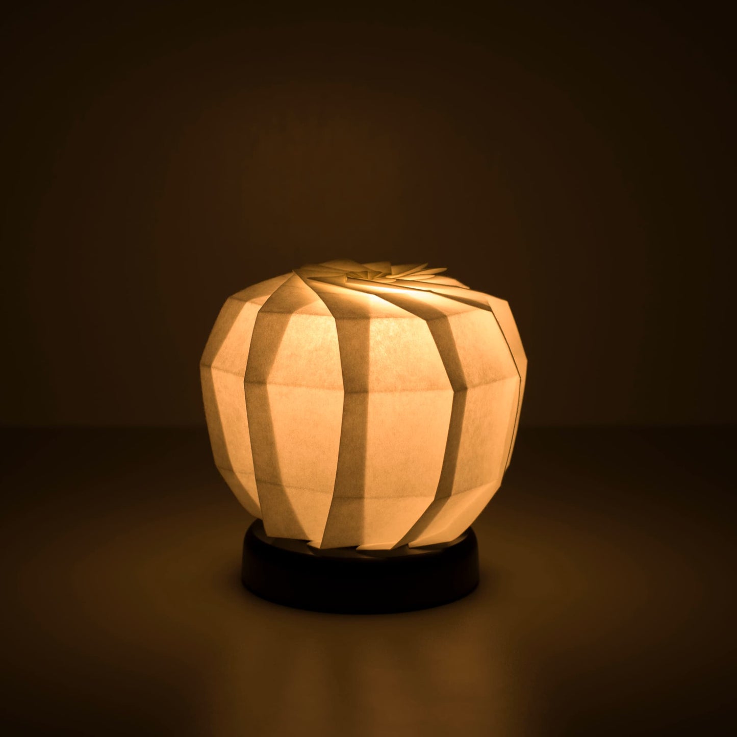 Origami papieren tafellamp "maru M" met een zwarte voet en een bolvormige witte lampenkap van ingewikkeld gevouwen washipapier, die een warm en uitnodigend licht uitstraalt.