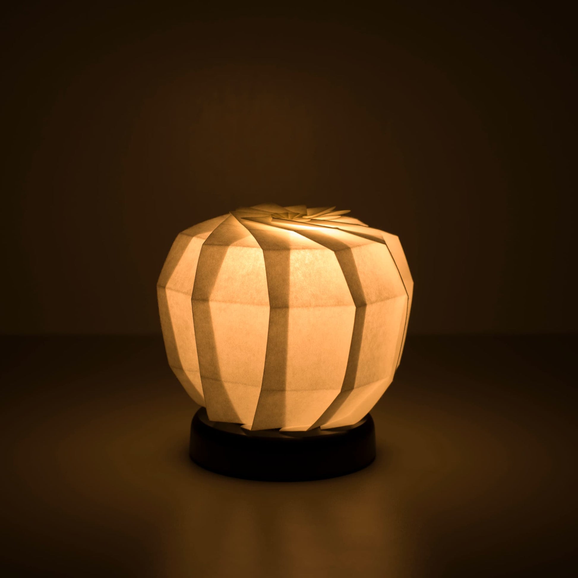 Origami papieren tafellamp "maru M" met een zwarte voet en een bolvormige witte lampenkap van ingewikkeld gevouwen washipapier, die een warm en uitnodigend licht uitstraalt.