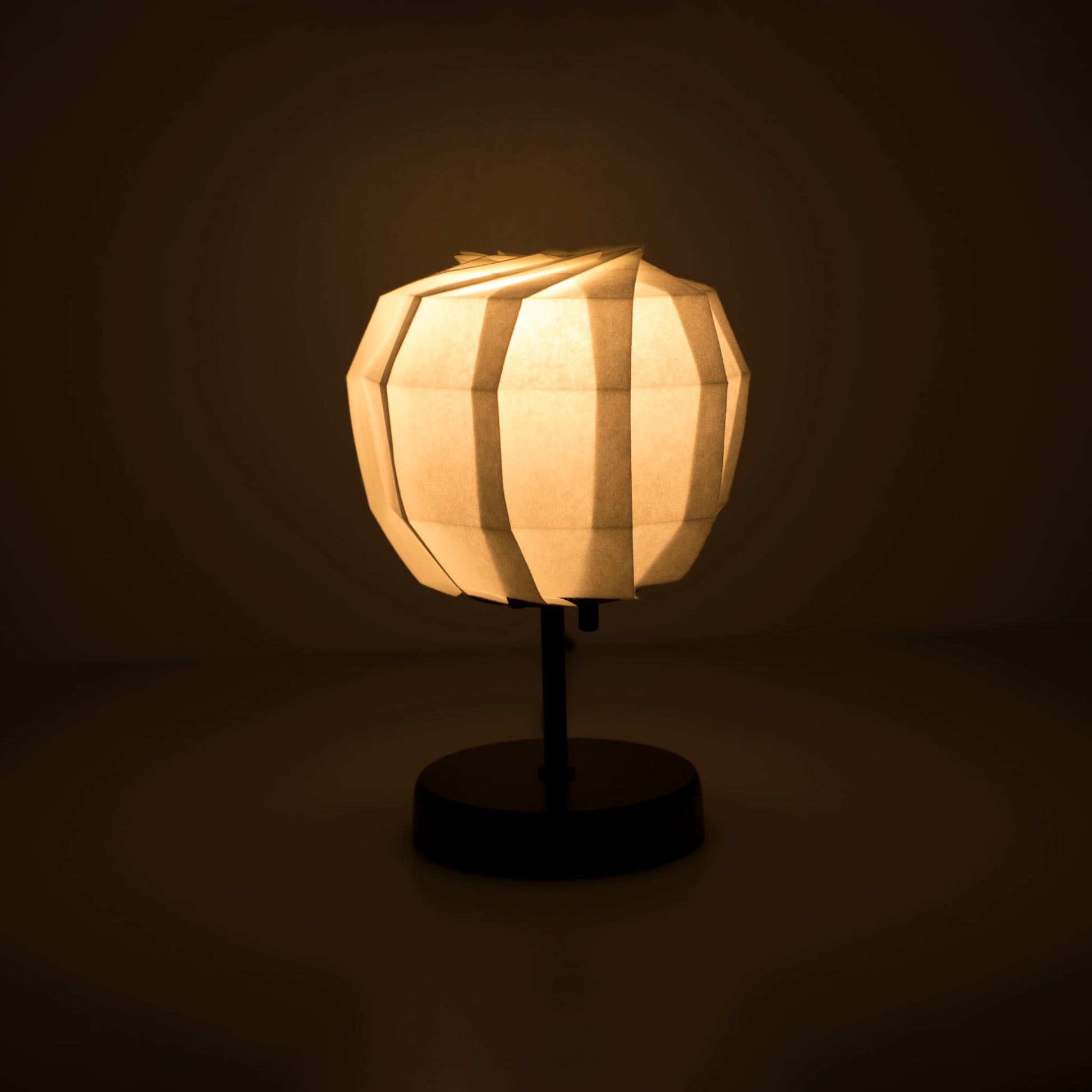 Origami papieren tafellamp "maru" met een zwarte standaard en een bolvormige witte lampenkap van ingewikkeld gevouwen washipapier, die een warm en uitnodigend licht uitstraalt.
