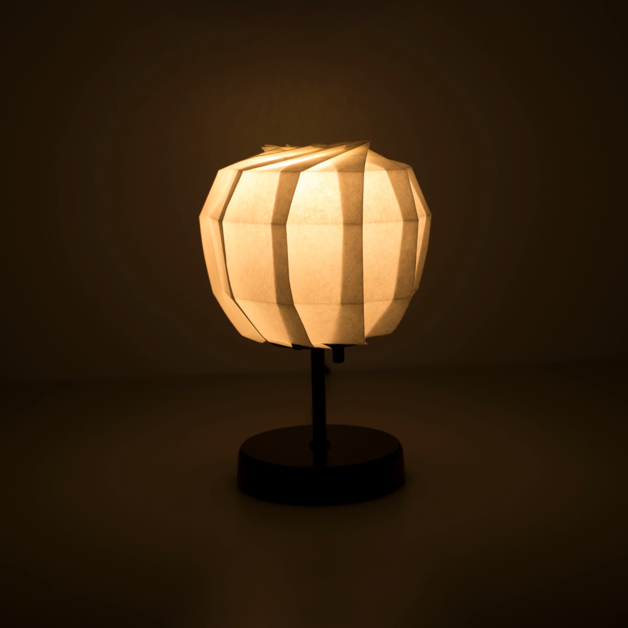 Origami papieren tafellamp "maru" met een zwarte standaard en een bolvormige witte lampenkap van ingewikkeld gevouwen washipapier, die een warm en uitnodigend licht uitstraalt.