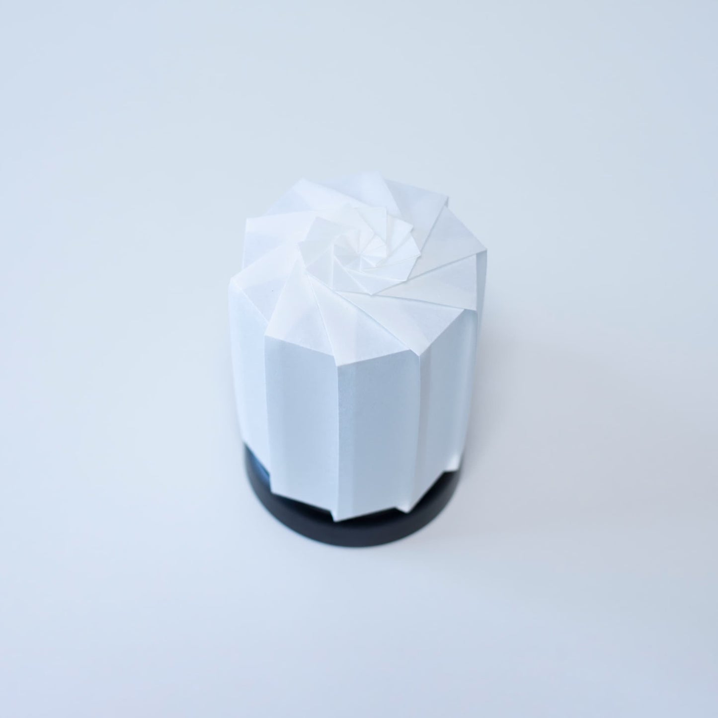 "Moderne Origami tafellamp tutu M" met een zwarte voet en een witte cilindrische lampenkap van getextureerd washipapier, wat zorgt voor een warme sfeer."