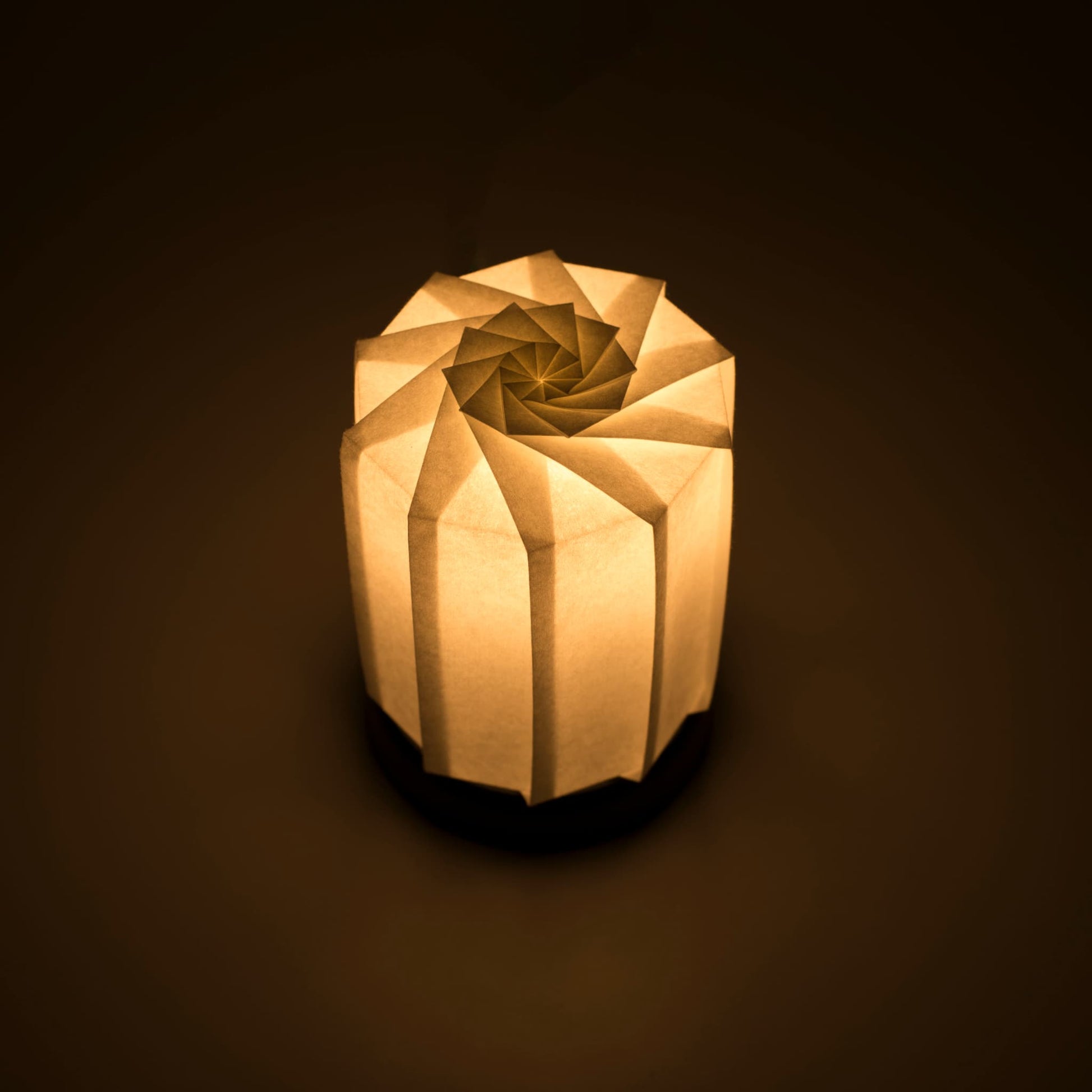 Origami papieren tafellamp "tutu M" met een zwarte voet en een bolvormige witte lampenkap gemaakt van ingewikkeld gevouwen washipapier, die zacht en warm licht uitstraalt.