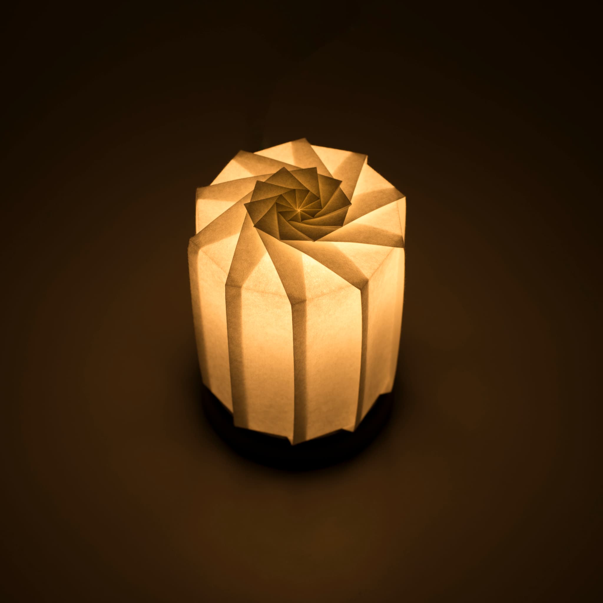 Origami papieren tafellamp "tutu M" met een zwarte voet en een bolvormige witte lampenkap gemaakt van ingewikkeld gevouwen washipapier, die zacht en warm licht uitstraalt.