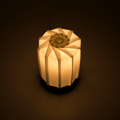 Origami papieren tafellamp "tutu M" met een zwarte voet en een bolvormige witte lampenkap gemaakt van ingewikkeld gevouwen washipapier, die zacht en warm licht uitstraalt.