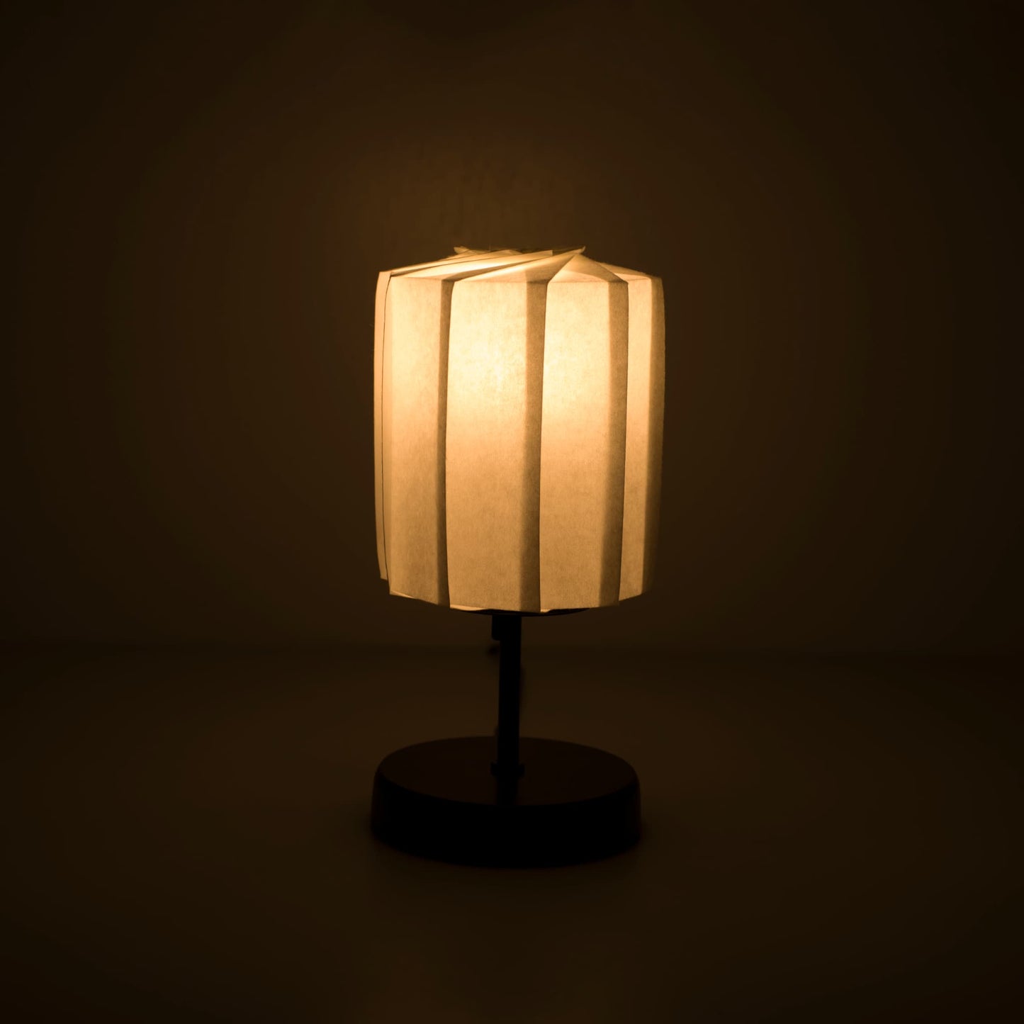 Origami papieren tafellamp "tutu" met een zwarte standaard en een cilindrische witte lampenkap van geplooid washipapier, die een zacht en warm licht werpt.