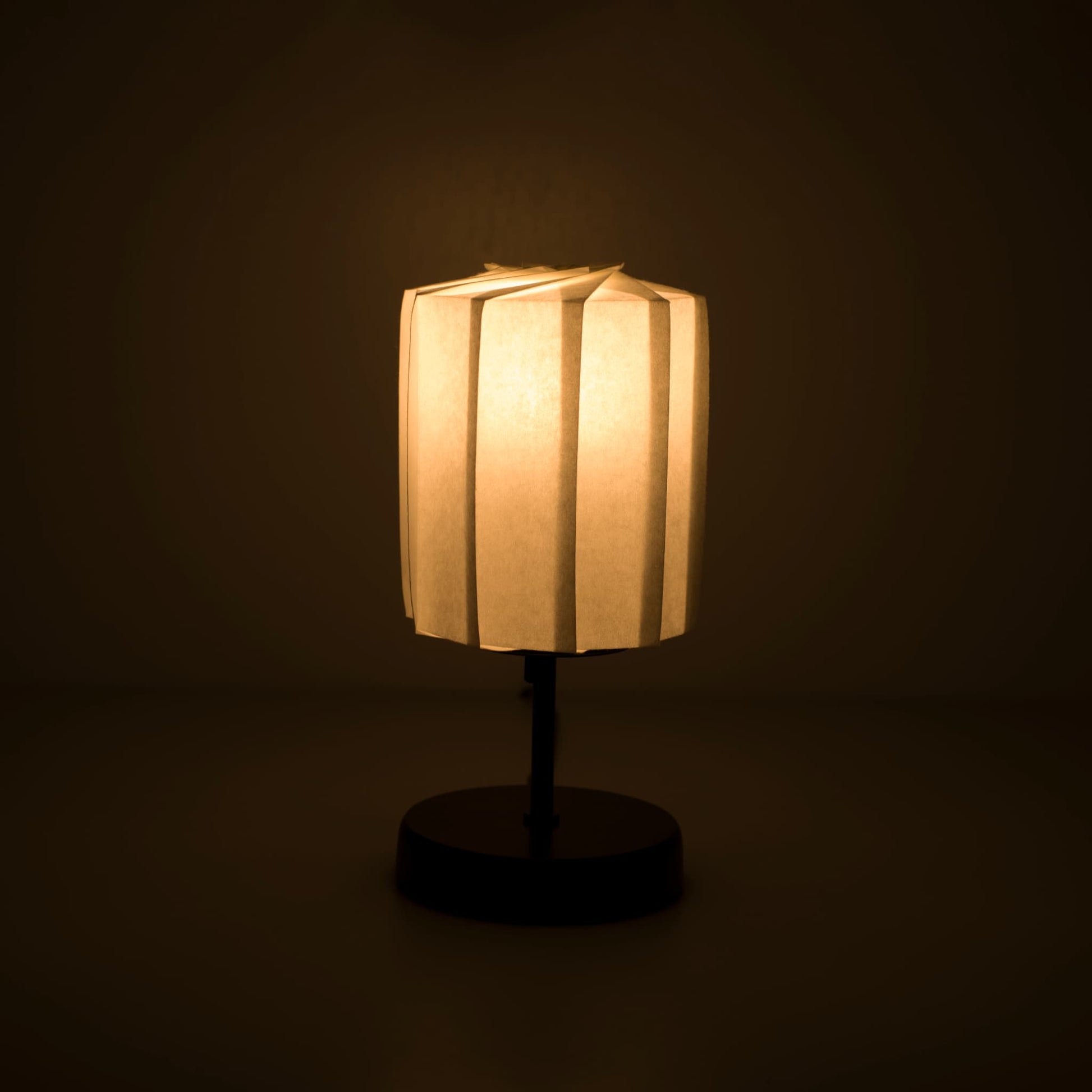 Origami papieren tafellamp "tutu" met een zwarte standaard en een cilindrische witte lampenkap van geplooid washipapier, die een zacht en warm licht werpt.