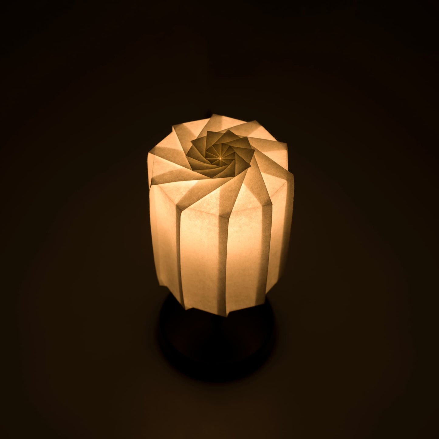 Origami papieren tafellamp "tutu" met een zwarte standaard en een cilindrische witte lampenkap van geplooid washipapir, die een warm en uitnodigend licht uitstraalt.