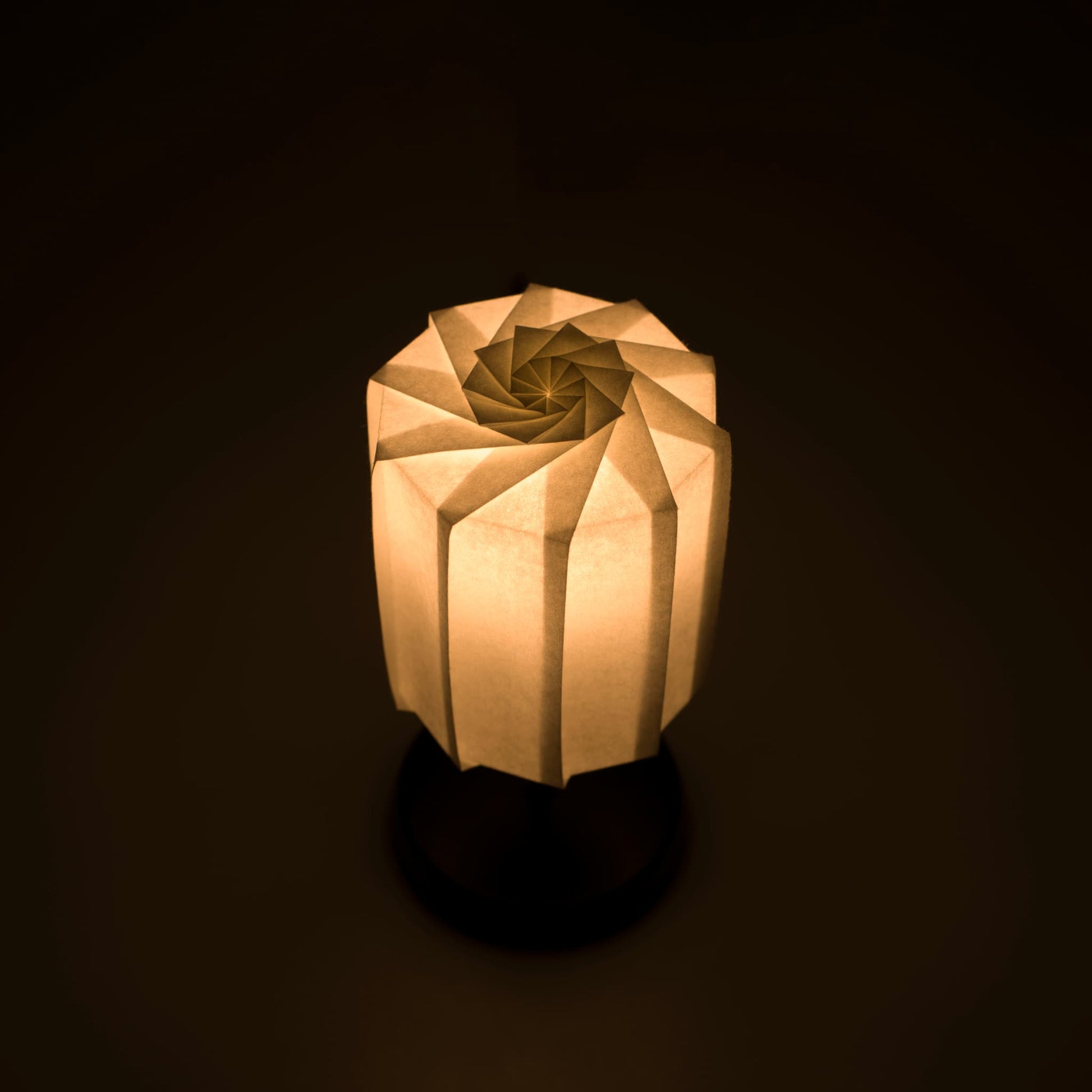 Origami papieren tafellamp "tutu" met een zwarte standaard en een cilindrische witte lampenkap van geplooid washipapir, die een warm en uitnodigend licht uitstraalt.