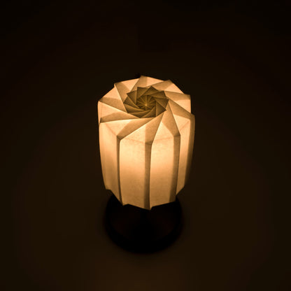 Origami papieren tafellamp "tutu" met een zwarte standaard en een cilindrische witte lampenkap van geplooid washipapir, die een warm en uitnodigend licht uitstraalt.
