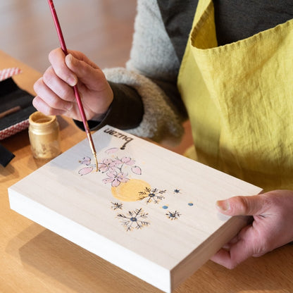 Tæt på en Buzan Kiln-håndværker, der maler kirsebærblomster og bladguld-design på en trækasse, hvilket udtrykker den raffinerede skønhed af traditionelt japansk håndværk.