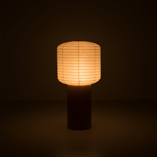 A portable Japanese paper lamp with a wooden handle and square-shaped cylindrical paper top emitting warm light in a dark room