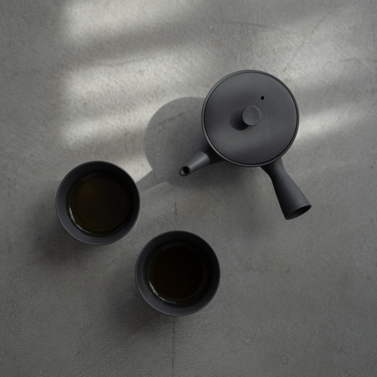 Schwarze Kyusu „Sencha“ und gefüllte japanische Teetassen auf einer Betonoberfläche für Minimalisten, die eine ruhige Teepause genießen.