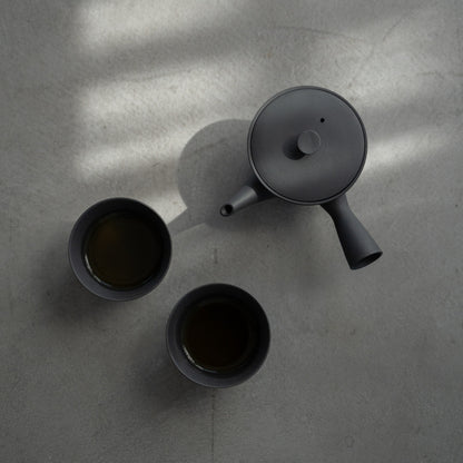 Schwarze Kyusu „Sencha“ und gefüllte japanische Teetassen auf einer Betonoberfläche für Minimalisten, die eine ruhige Teepause genießen.