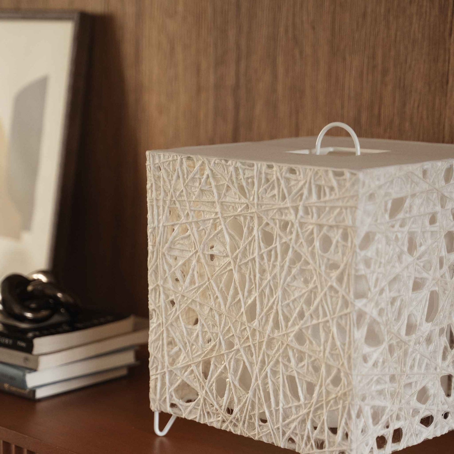 "Lampe japonaise blanche en forme de cube en papier Me Look 03, posée éteinte sur une console en bois près d'un arrière-plan de lit."