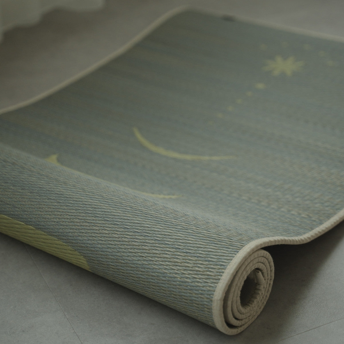 Grijze Japanse Tatami Yogamat "Maan" gedeeltelijk opgerold op een vloer in betonlook, met de fijne, authentieke textuur van geweven natuurlijk matgras.