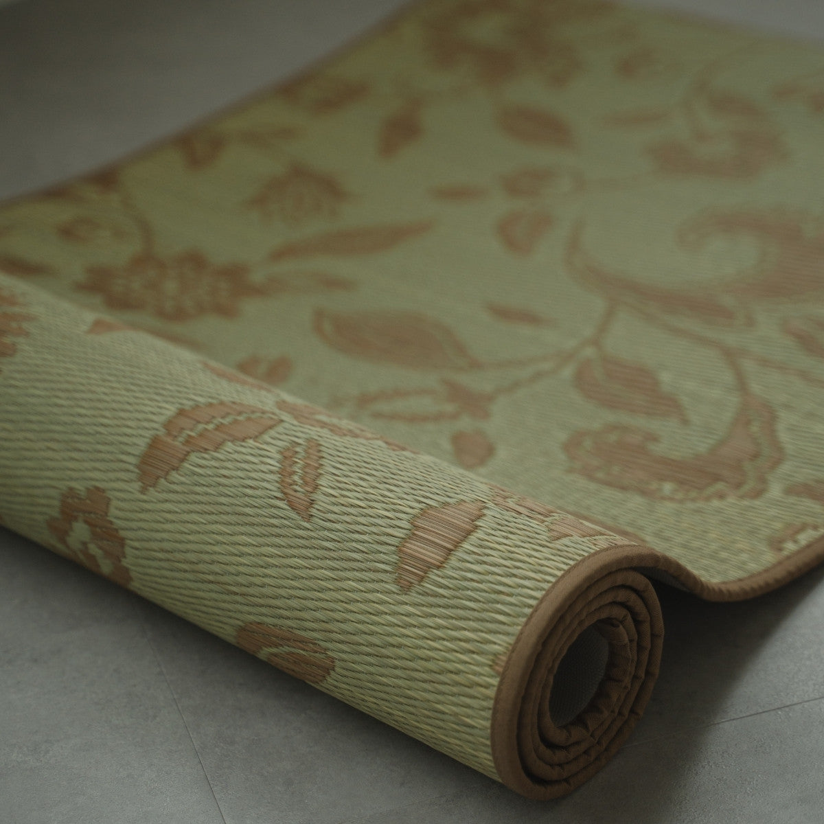 Close-up of Japanese Tatami Yoga Mattress "Sarasa" Beige partially rolled on a grey floor, showing woven natural rush grass texture.