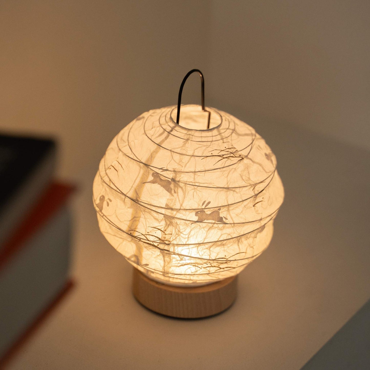 Glowing round Japanese paper table lamp "Yuhi" Rabbit serving as a cozy accent lamp next to books on a white console table.