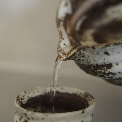 Sake pouring from a textured Zui Japanese sake carafe into a white ceramic sake cup, to effortlessly enjoy warm sake