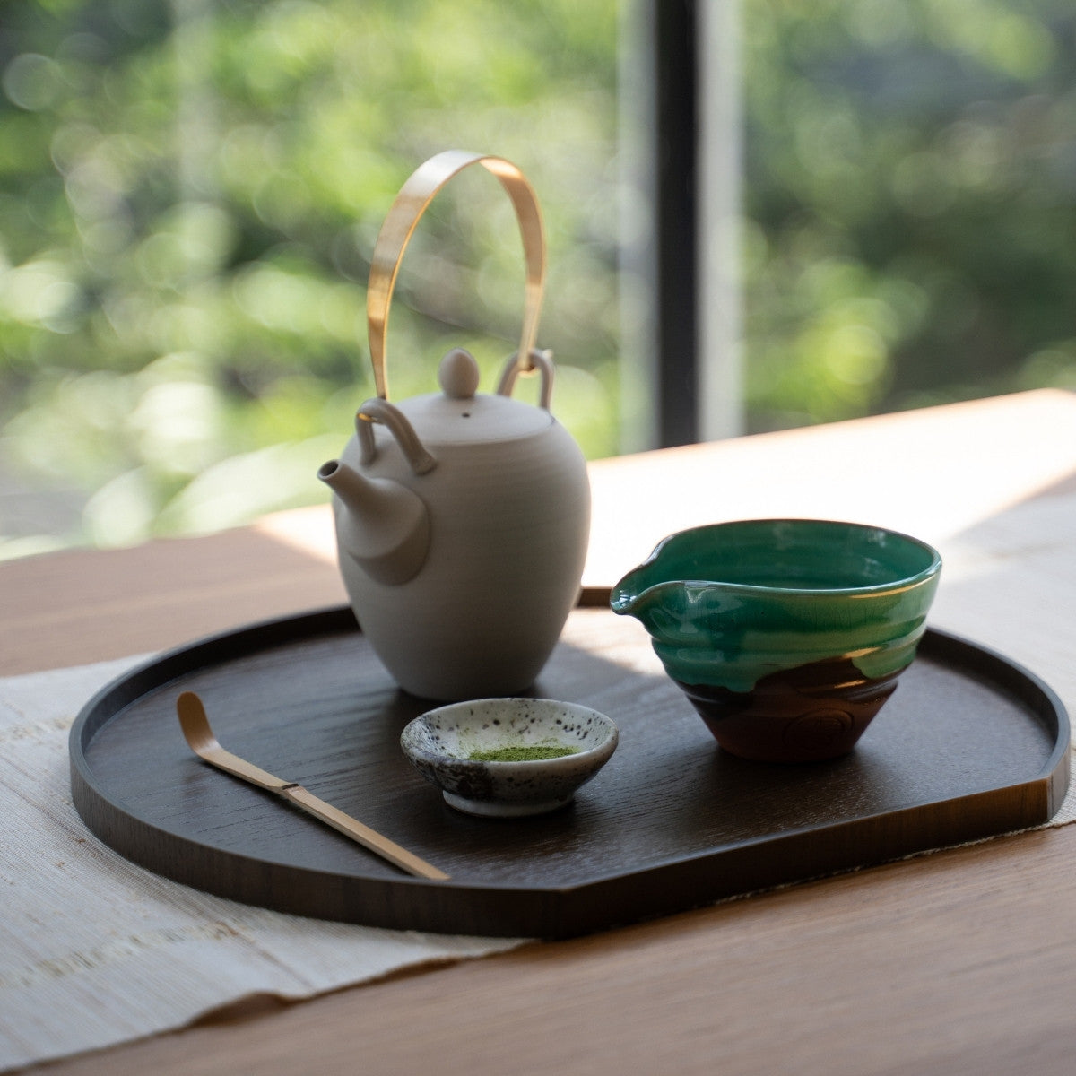 Smaragdgrøn japansk matcha-skål med tud og te-redskaber på en mørk bakke ved et lyst vindue, klar til at lave lattes til gæster.
