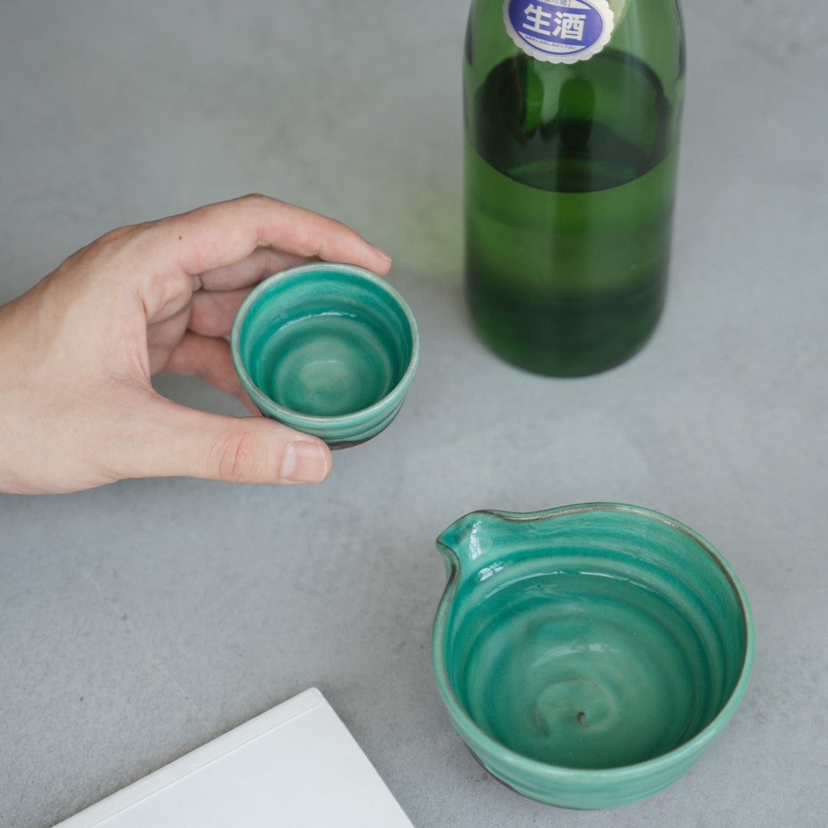 A hand holds an emerald Japanese sake cup over a grey table, with a matching sake carafe ready for a mature evening drink.