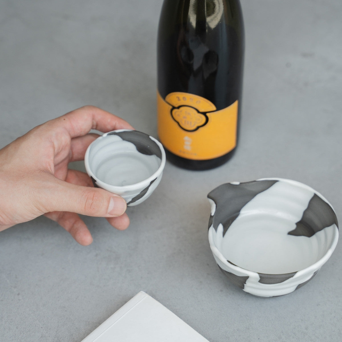 A hand holds a White Flow Japanese sake cup by a carafe and book on a grey table, for a relaxing evening sake ritual