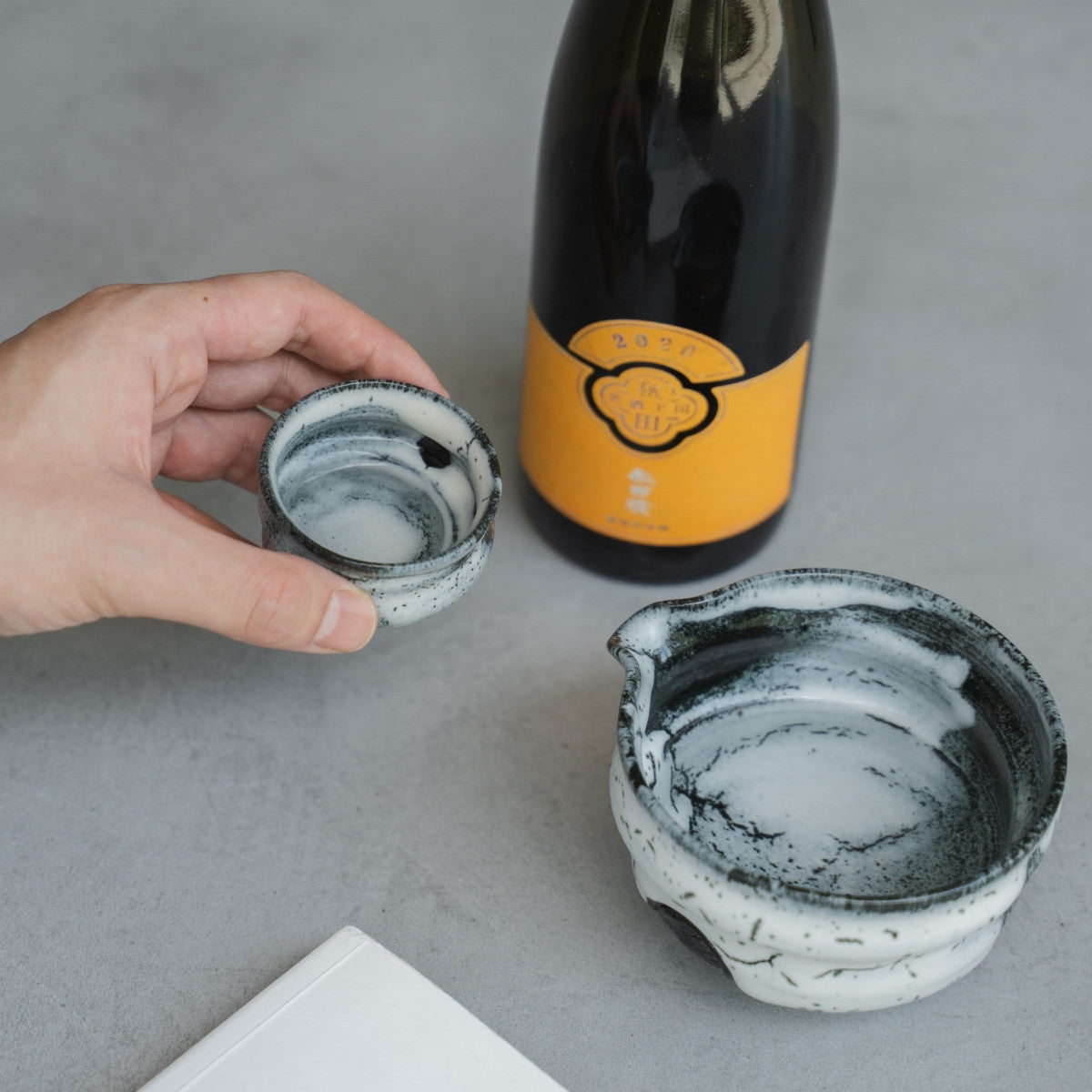 A hand holds a Creative Cloud Japanese sake cup next to a sake carafe and book, for a relaxing wabi-sabi evening