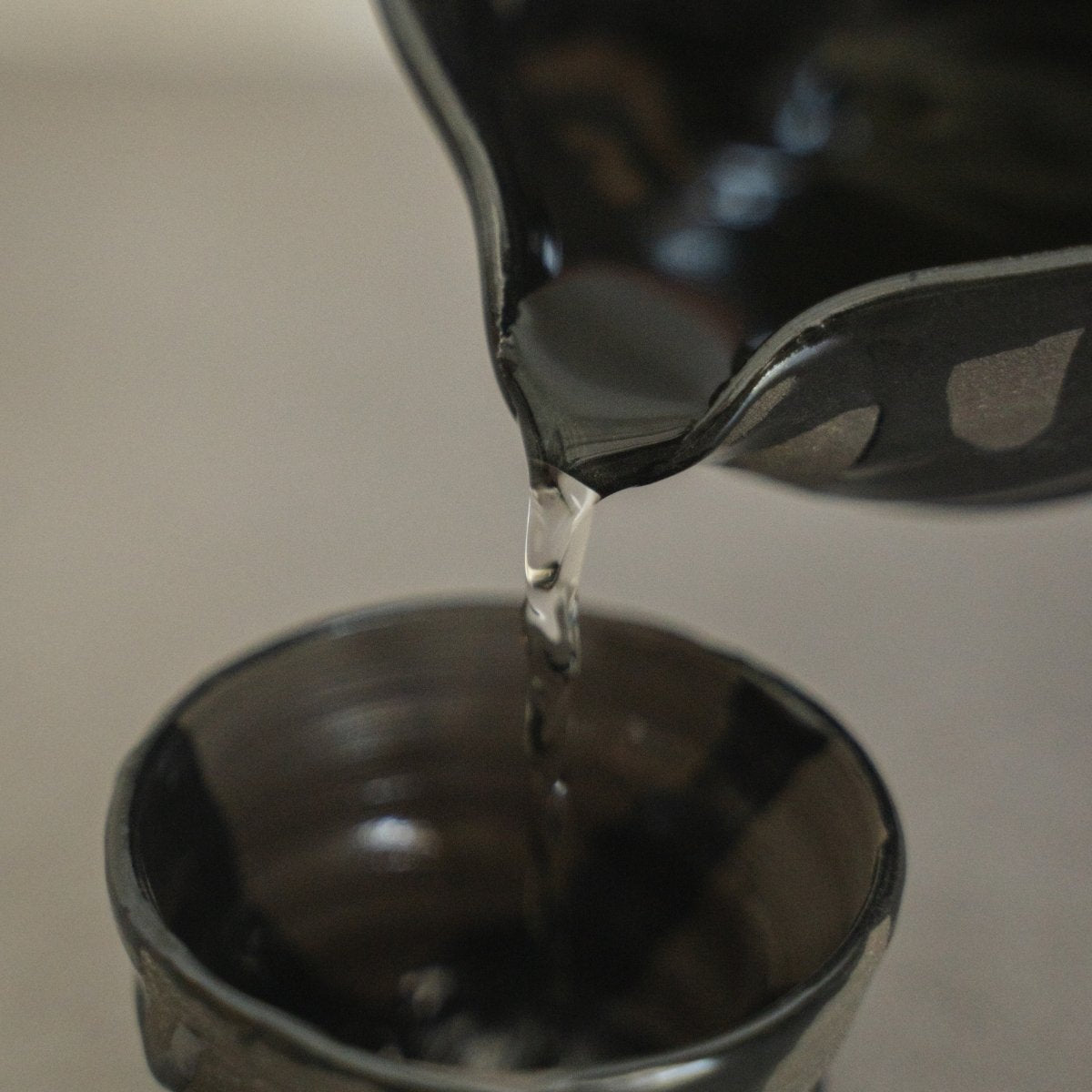 Nærbillede af væske, der hældes fra tudet på en Sumi Black japansk sake-karaffel, ned i en håndkastet sakekop, til bevidst nydelse af varm sake.