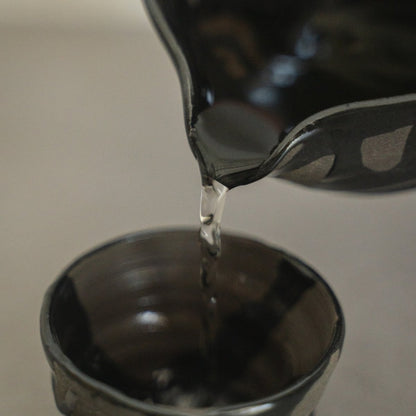 Nærbillede af væske, der hældes fra tudet på en Sumi Black japansk sake-karaffel, ned i en håndkastet sakekop, til bevidst nydelse af varm sake.