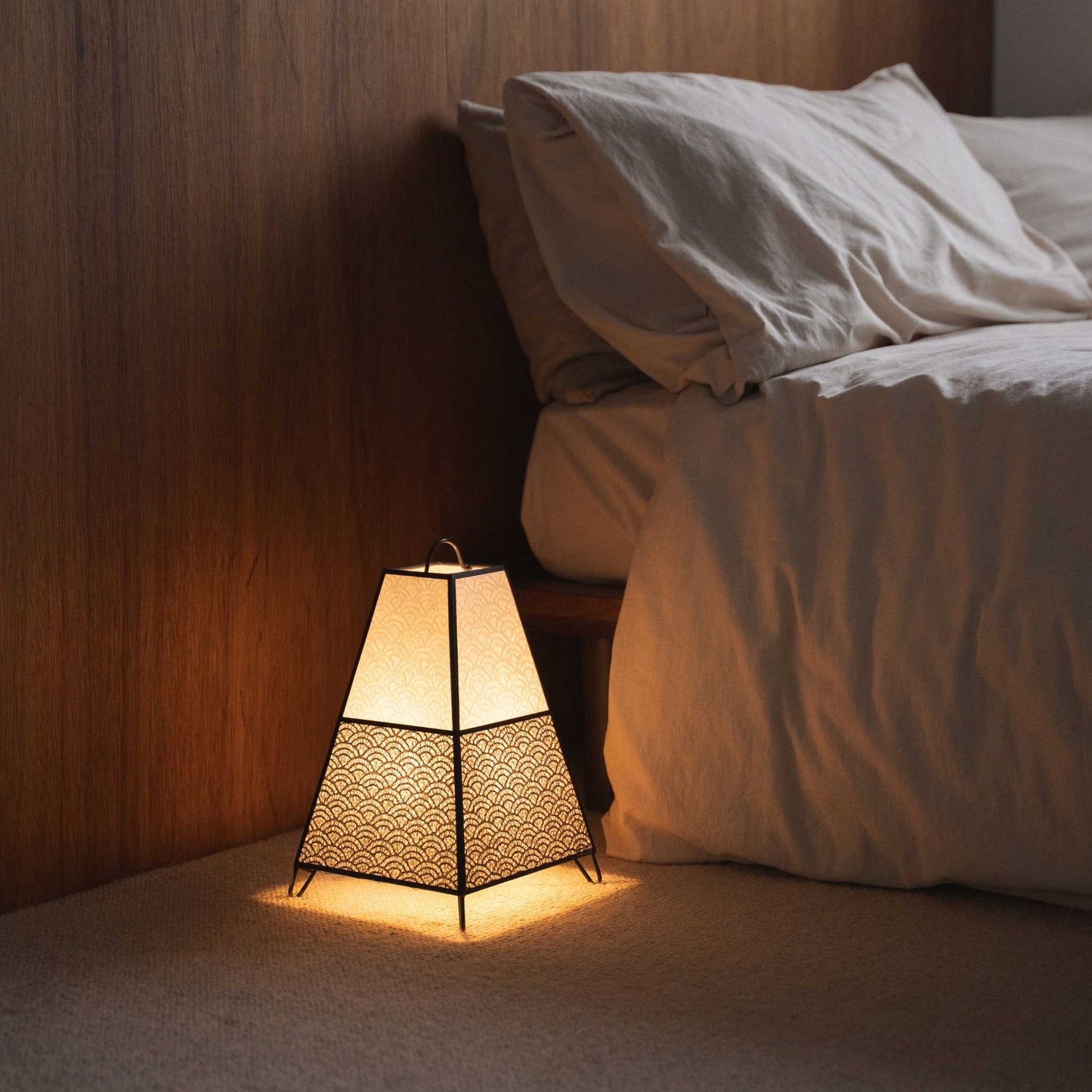"Lit Japanse vloerlamp KURA Small werpt ingewikkelde schaduwen van zijn papieren kap als sfeerverlichting in een Japandi slaapkamer."