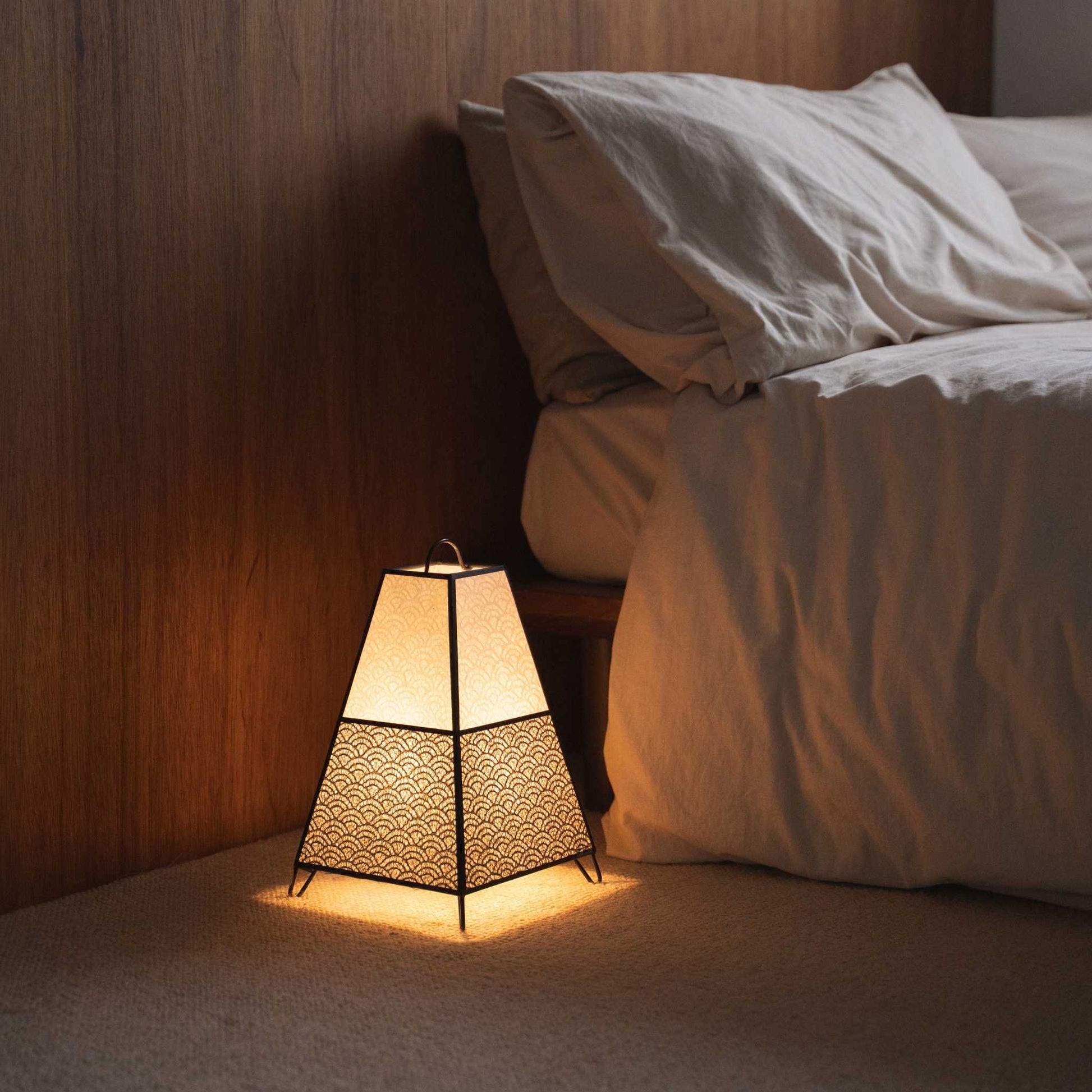 "Lit Japanse vloerlamp KURA Small werpt ingewikkelde schaduwen van zijn papieren kap als sfeerverlichting in een Japandi slaapkamer."