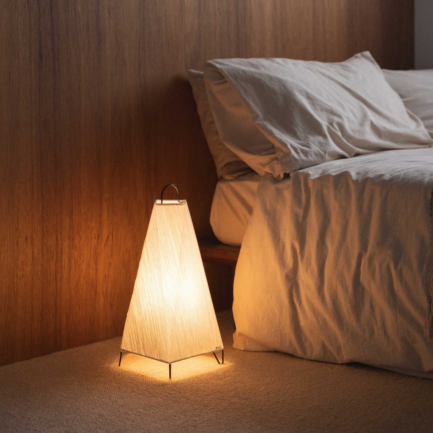 Lit japansk papir gulvlampe ORI lille kaster et varmt skær som en hyggelig ambient lampe ved siden af en seng i et afslappende soveværelse.
