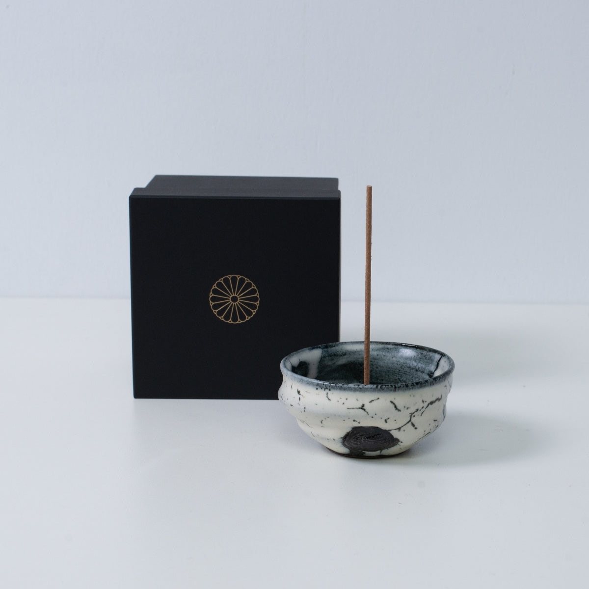 White and black Japanese incense holder "Creative Cloud" bowl with a stick next to a black gift box with a gold chrysanthemum crest, Japan.