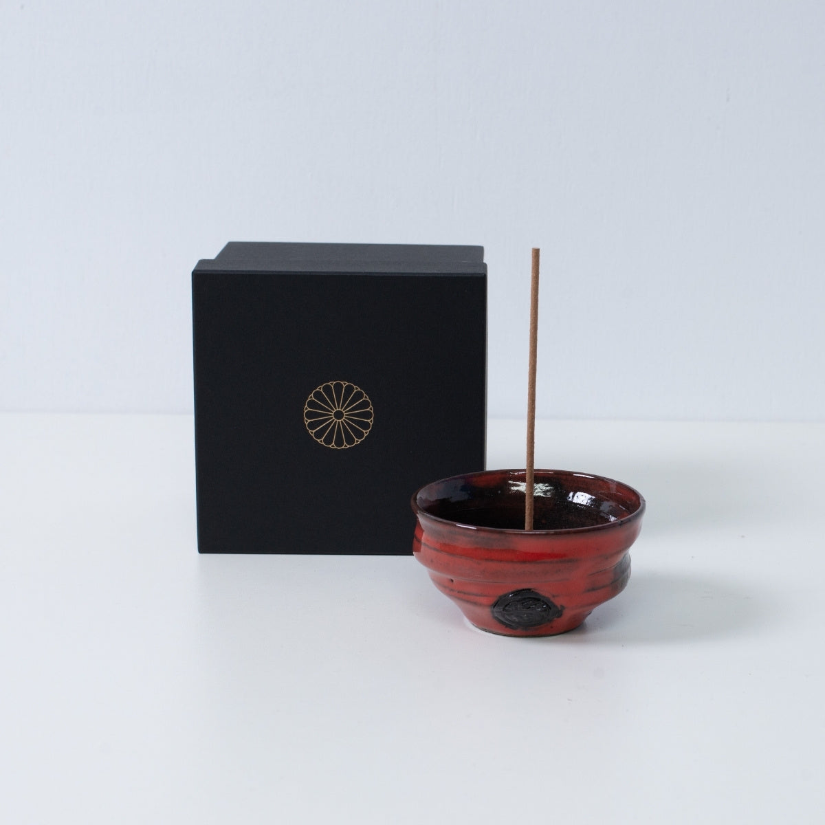 Crimson og sort japansk røgelsesholder "Nobel Red" keramikskål med en pind ved siden af en sort gaveæske med et gyldent krysantemum-emblem, Japan.