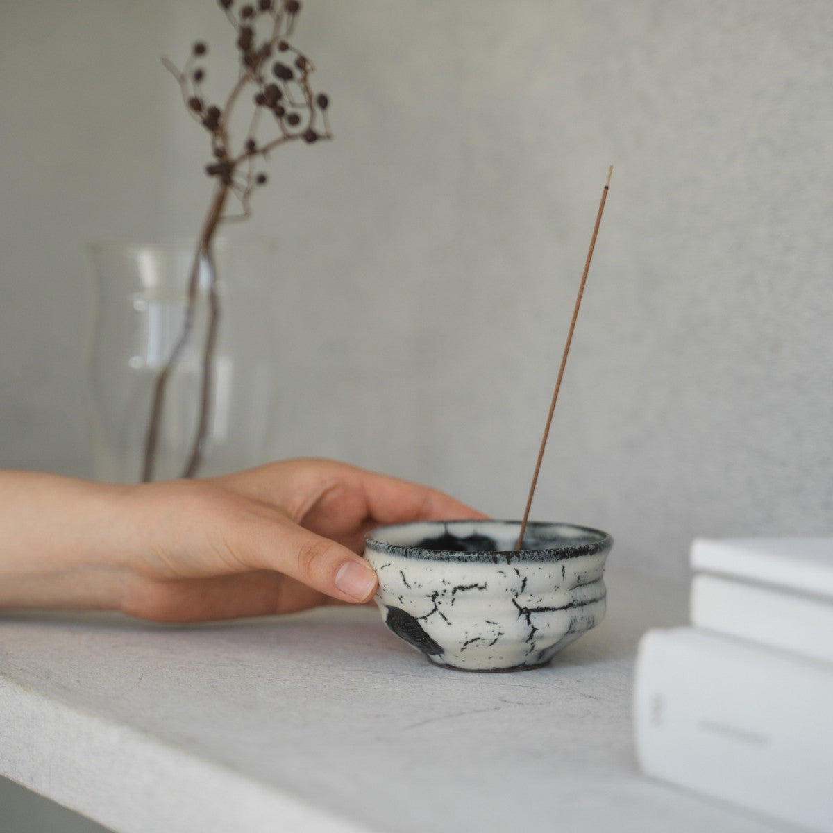 A hand reaching for a white ceramic Japanese incense burner on a bright shelf, preparing for a calming meditation ritual.